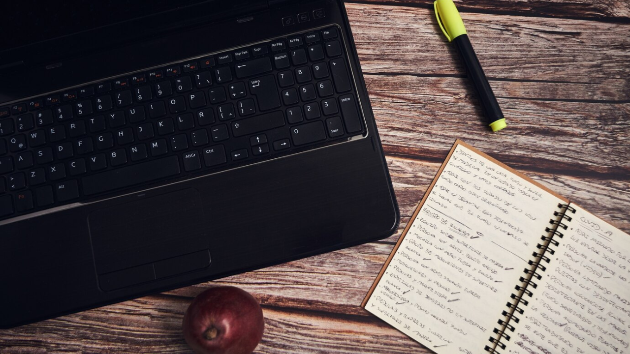 Laptop, spiral notebook filled with writing, a marker, and an apple on a wooden table.